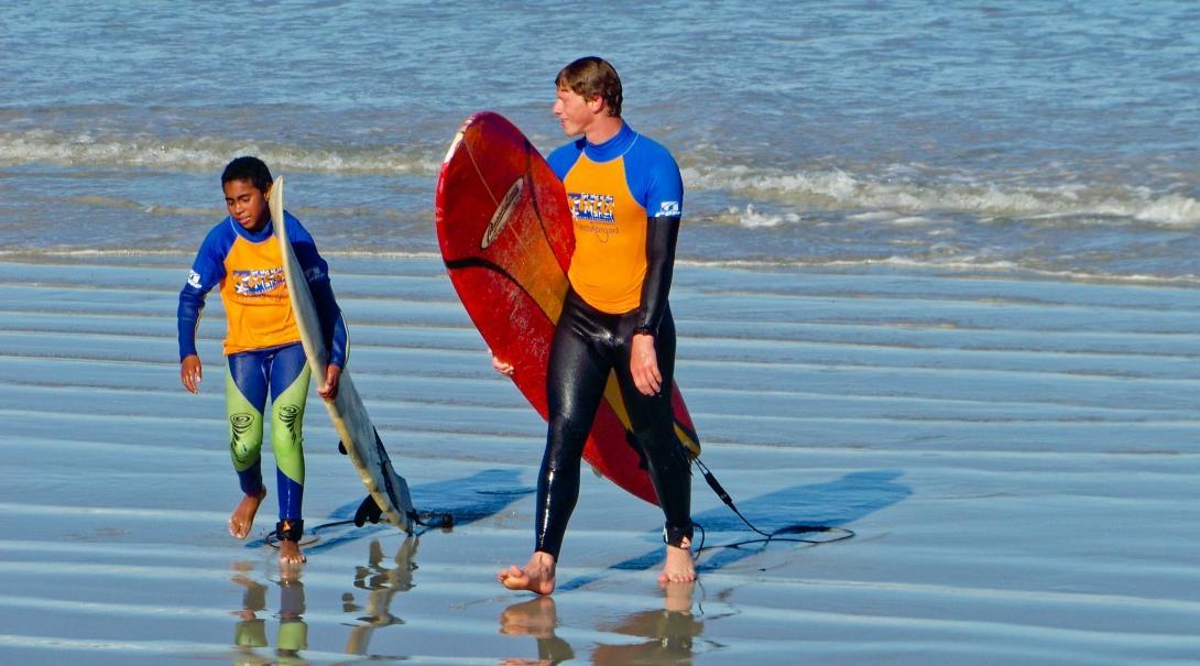 A Projects Abroad volunteer teaching children to surf in South Africa walks along the beach with a child A volunteer works closely with a child to teach surfing in South Africa